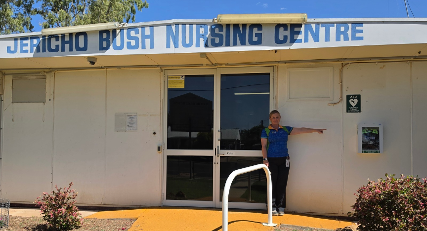 A woman in blue shirt stands in front of a single-level building pointing at a small square device on the wall.