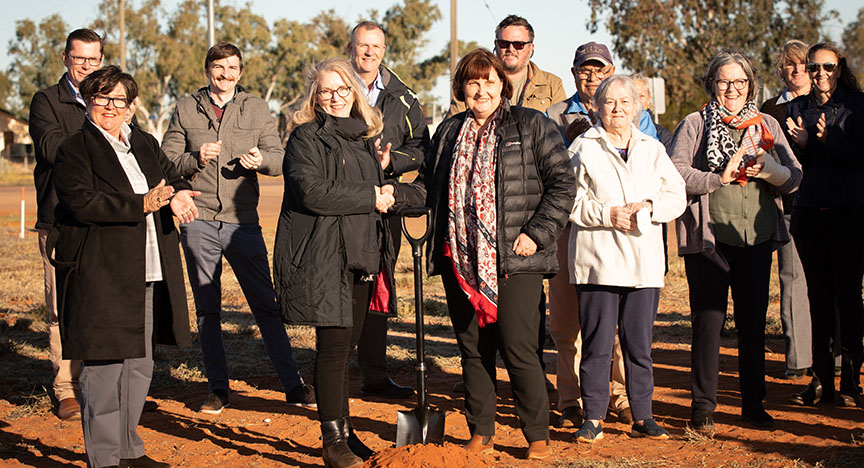A group of people stand in red soil facing the sun. Some of the people hold spades.