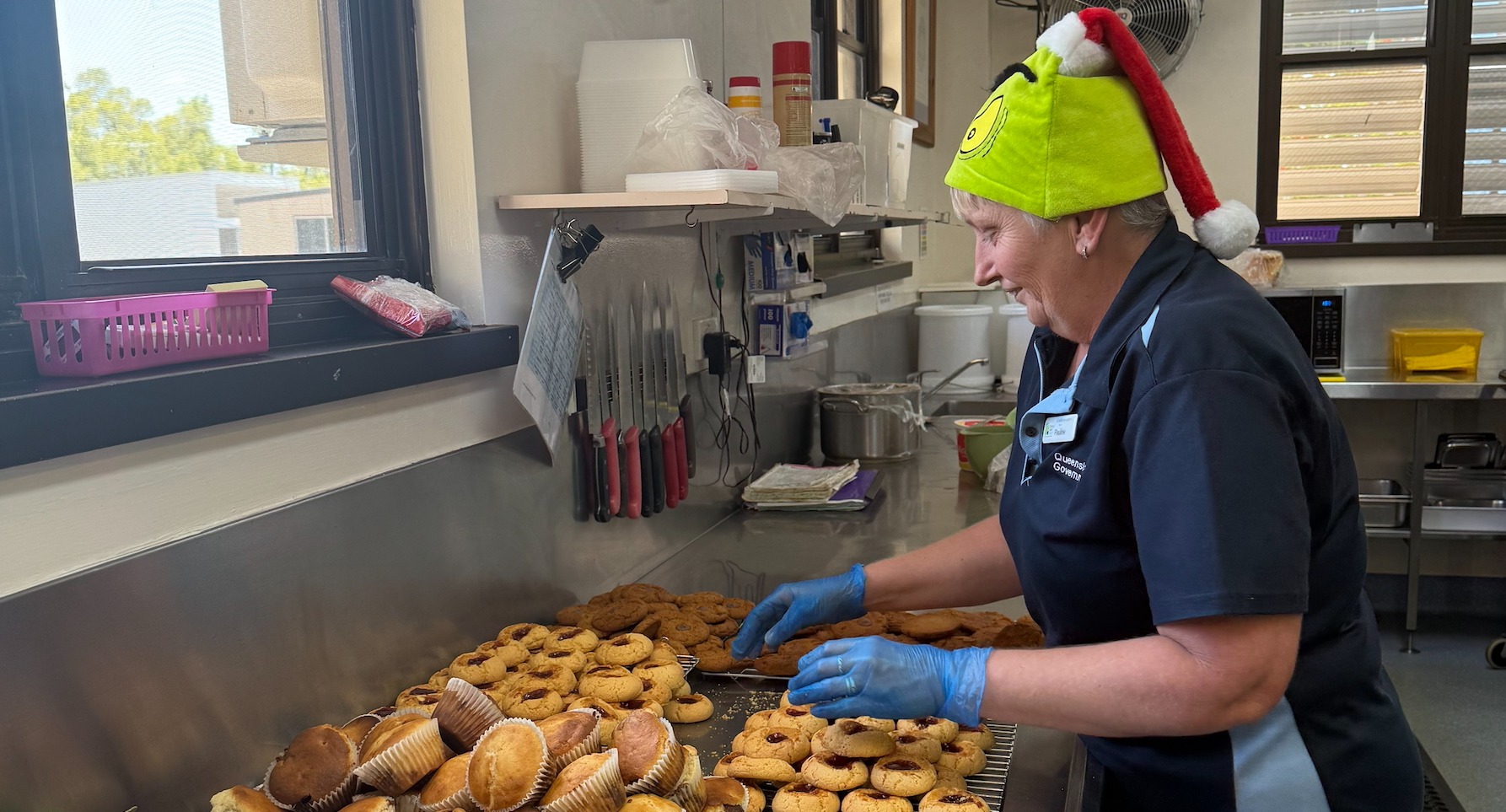 A female cook with a green Grinch-themed santa hat prepares cookies and muffins on a steel tray.