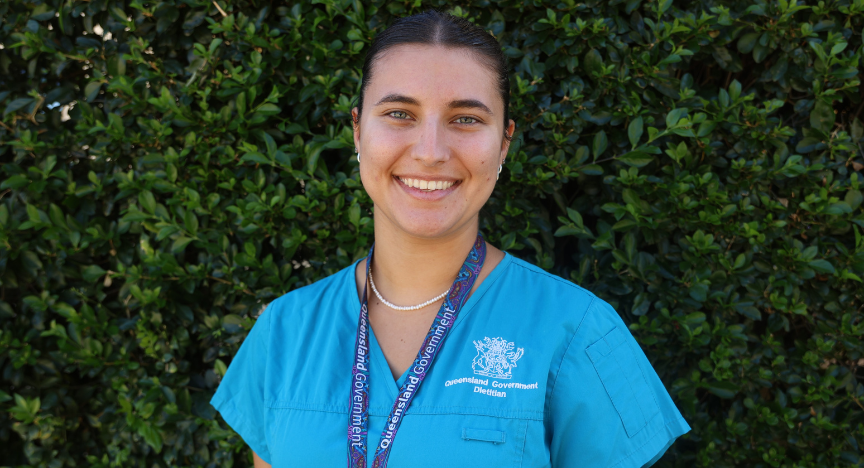 A lady with dark hair in blue scrubs smiles at the camera in front of a tall green hedge.