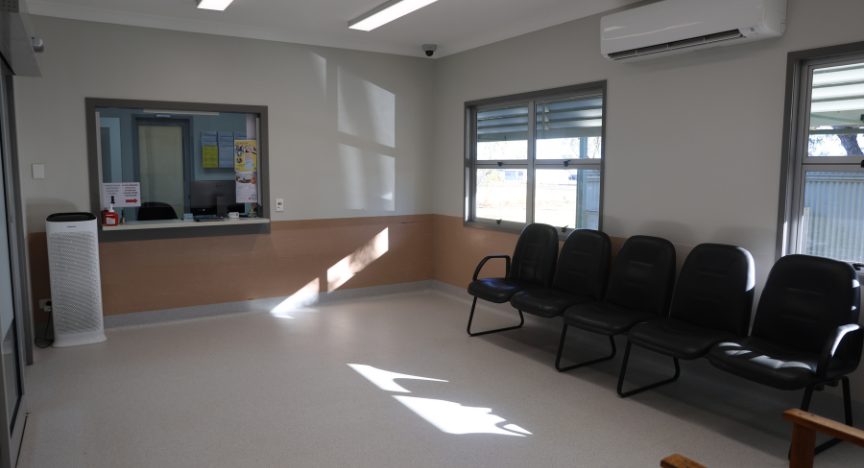 The interior shot of a waiting room, featuring white floors and walls, dark chairs, and an admin window.