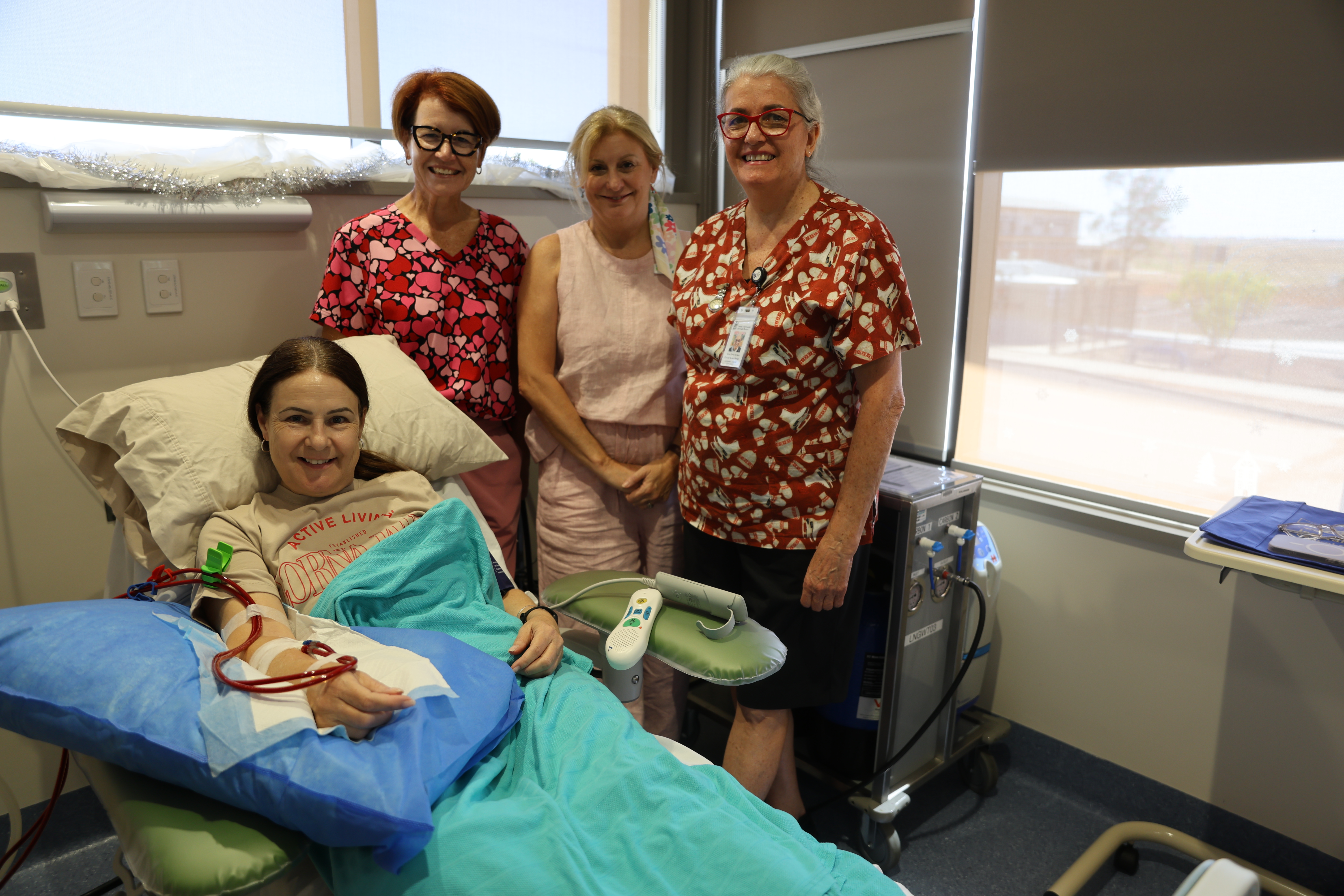 A group of four people smile at the camera in a hospital room, with one dark haired woman laying in a bed receiving dialysis.