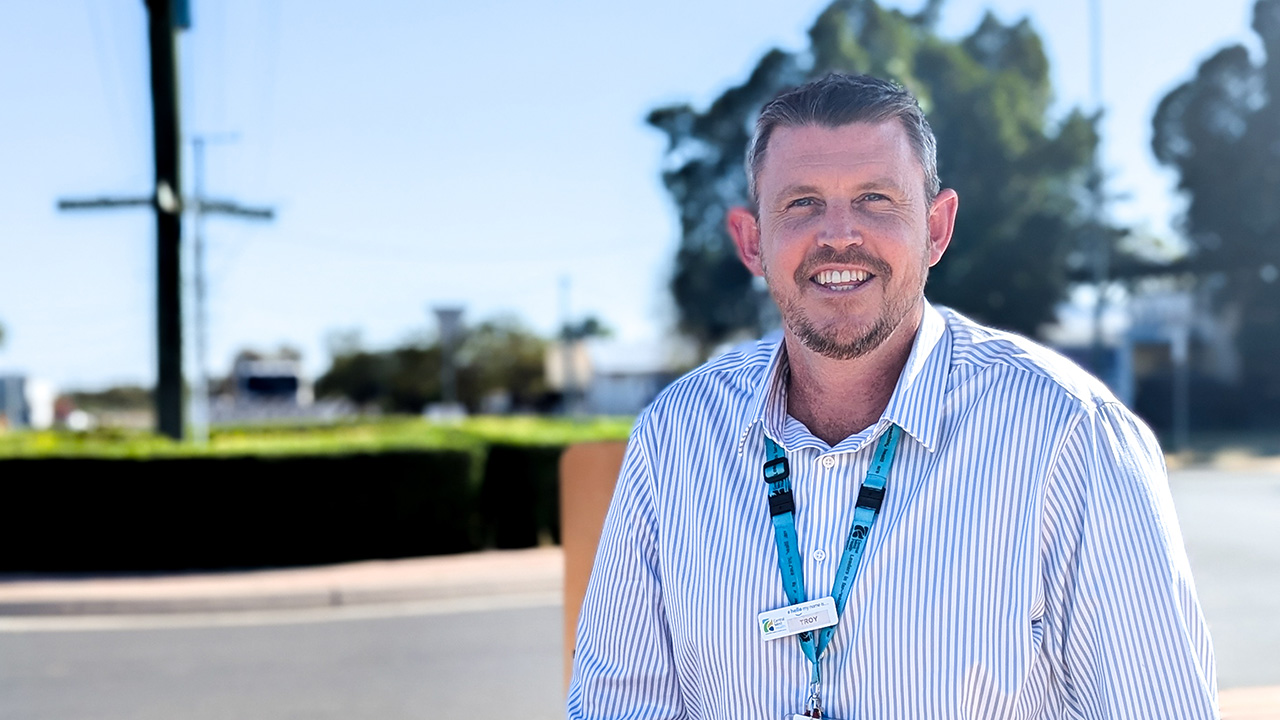 A man with short dark hair and a light beard in a blue-striped shirt smiles at the camera with the backdrop of a rural town.