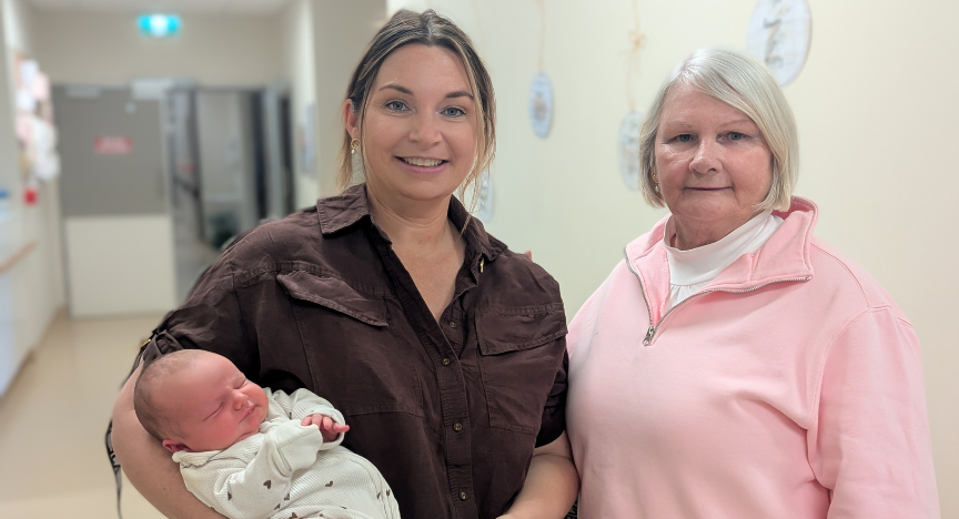 A lady in a brown shirt holds a newborn baby, as she stands next to her mother in a hospital hallway.