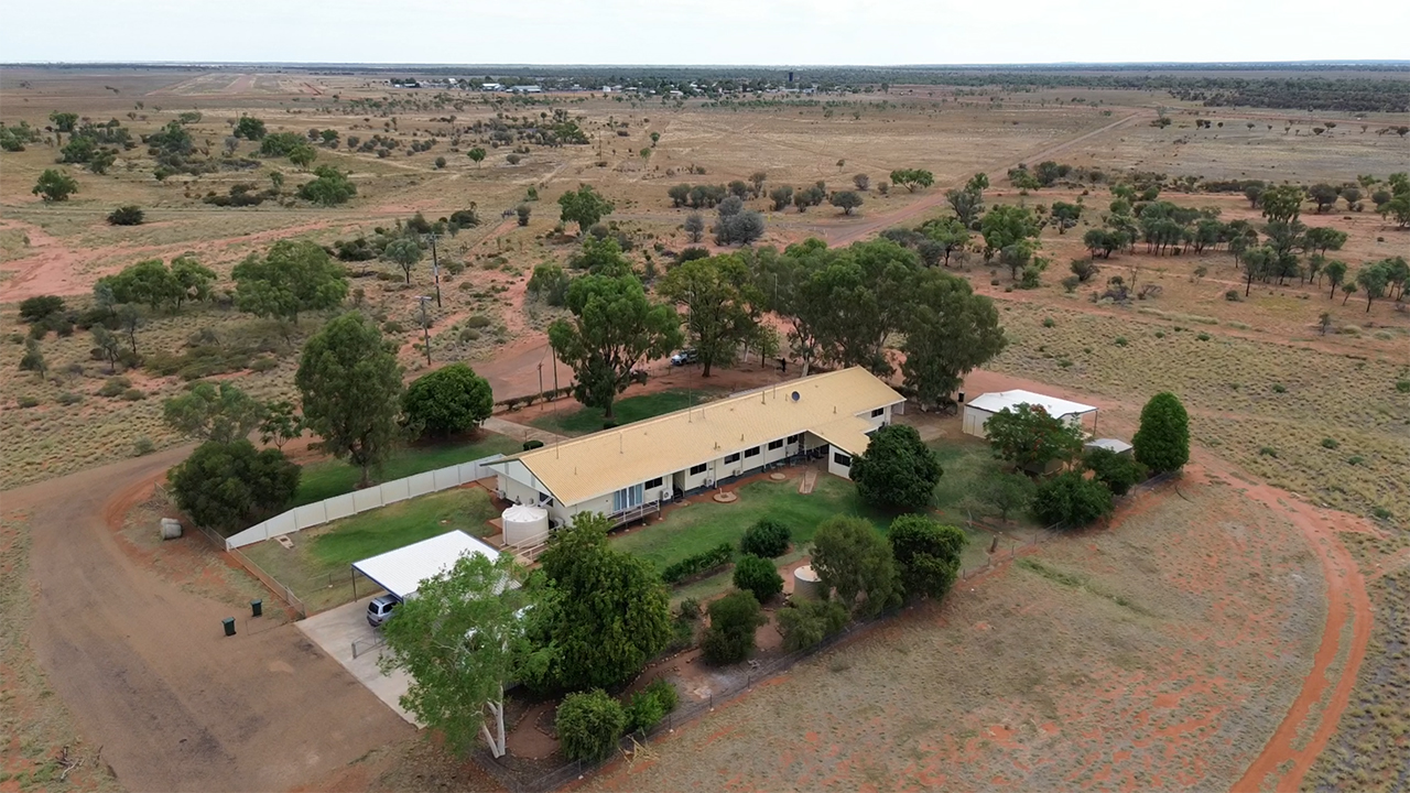 An aerial image of a small, long building surrounded by green in its immediate vicinity, then drier landscape all the way to a small town in the distance.