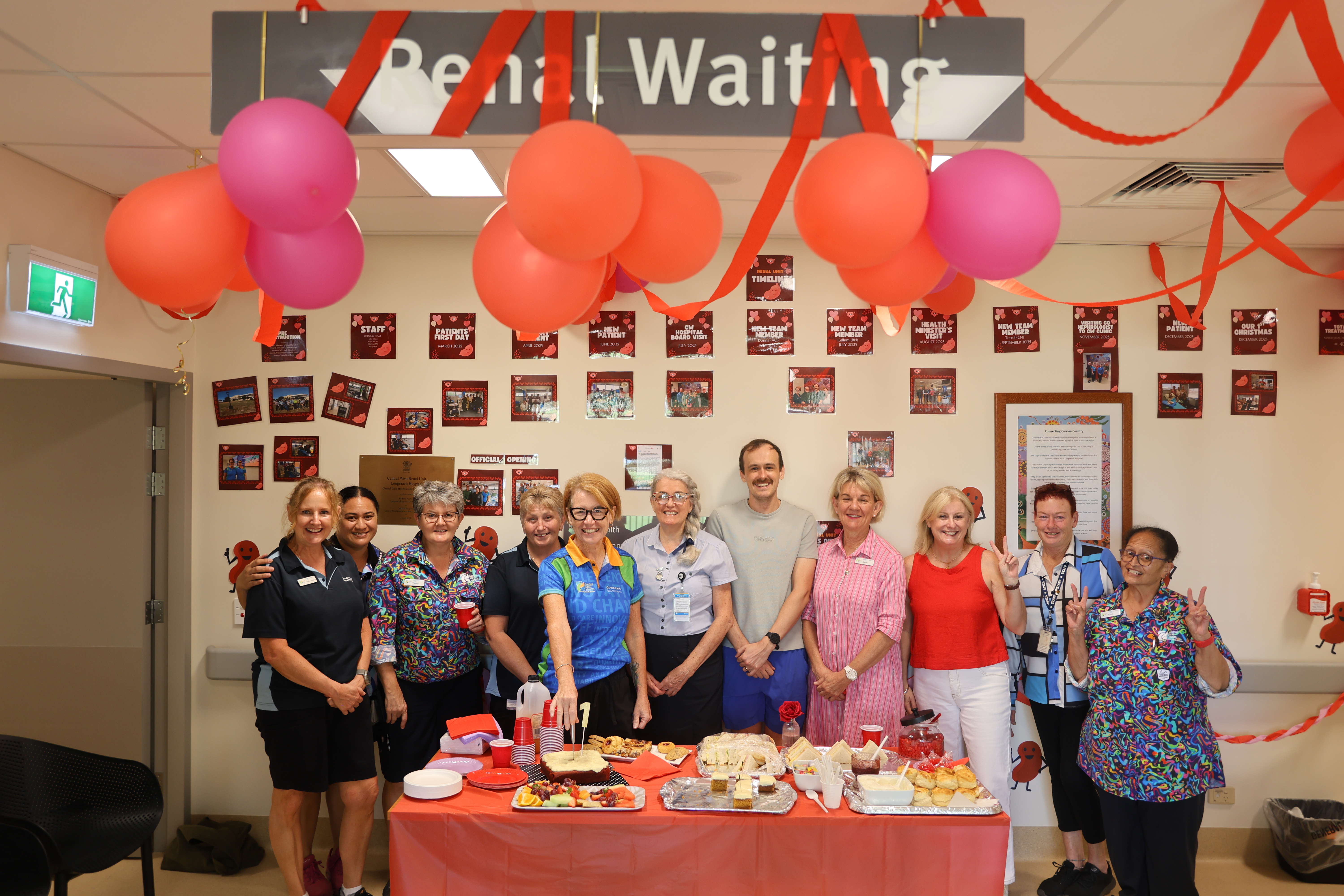 A group of people smile for the camera in a room decorated with red balloons, streamers, and a table full of party food.