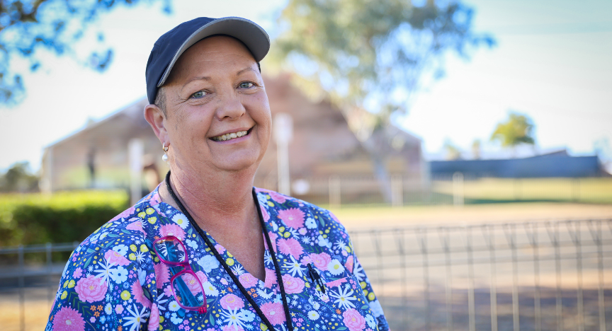 A woman wearing a cap and a colourful shirt smiles at the camera while standing outside.